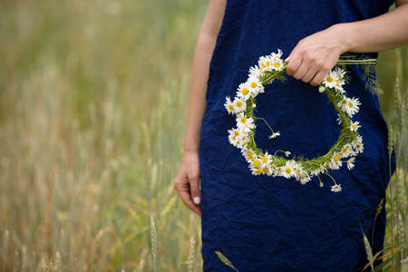 Closeup of young woman's hands holding wreath with wild flowers while walking on wheat or rye field on a summer day. Countryside landscape. Girl with chamomiles.の写真素材
