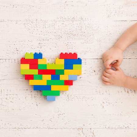 Top  view  on child's hands playing with bright heart made of  colorful plastic bricks on old wooden...