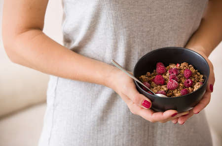 Closeup of girl's hands holding black bowl with organic granola with coconut milk and raspberries.の写真素材