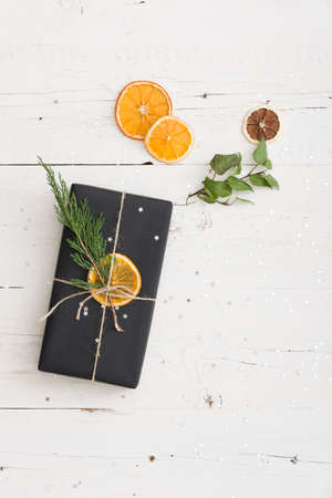 Top view on nice Christmas or birthday gift wrapped in black paper and decorated with cord, orange chips and tree brunch on white wooden background.の写真素材