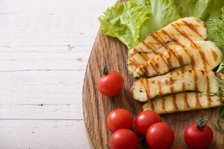 Grilled slices of homemade halloumi cheese with green salad, fresh herbs and organic tomatoes. Fried halloumi cheese with grill marks on wooden background, top view, close-upの写真素材