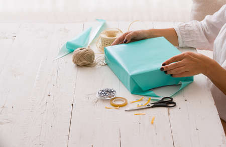 Closeup of woman's hands with gift ribbon, twine, tape and present wrapped in turquoise gift paper on white wooden background. Christmas, birthday or any other celebration preparations.の写真素材