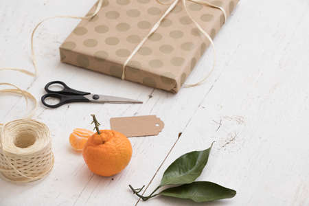 Close up of gift wrapped in craft dotted paper, card, scissors, ribbons, tangerine and tag on white wooden background. Holidays season.の写真素材