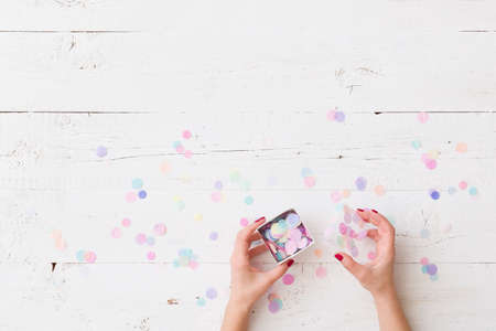 Top view on girl's hands holding small bow with tissue paper confetti on white wooden table background. Holiday season. Celebration.の写真素材