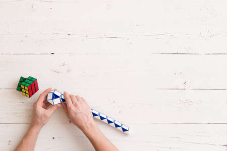 Moscow, Russia, February 16 2018: Top view on Rubik's snake puzzle in man's hands, and Rubik's cubes laying on white wooden table or background. Man holding Rubik's puzzle toy and playing with it.の写真素材