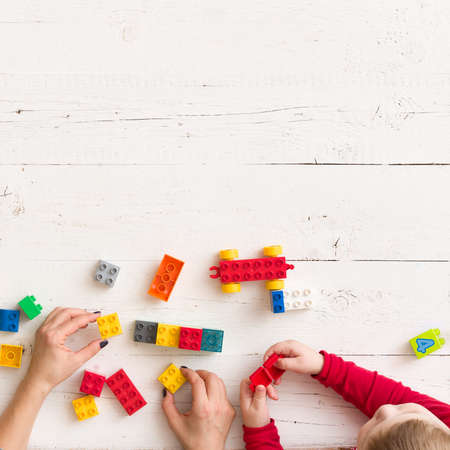 Closeup of young woman's and child's hands playing with toys and plastic bricks on white wooden table backgroundの写真素材