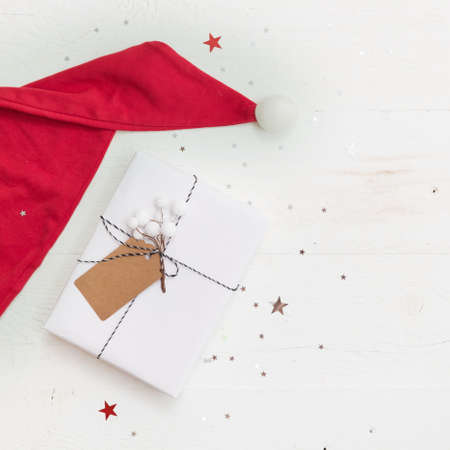 Santa's red hat and nice Christmas gifts box wrapped in white paper and decorated with ribbon and tag on old white wooden background with shinny silver stars. Christmas and holidays season concept.の写真素材