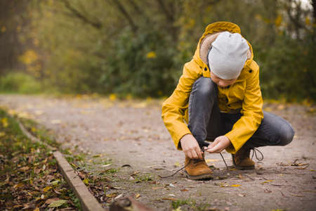 Cute child tying laces while walking in the city park on a cold autumn day. Kid boy outdorsの写真素材