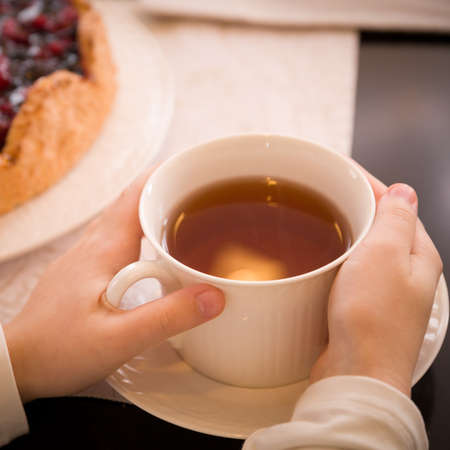 Closeup of a young girl's hand holding a beautiful white cup with tea on the background of a birthday cake. Celebration conceptの写真素材