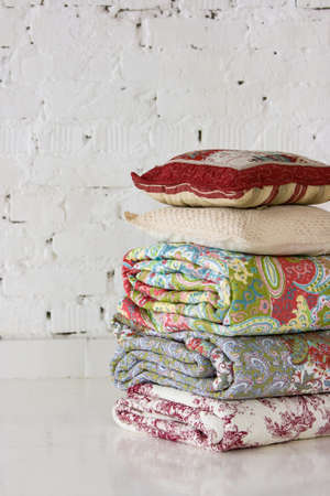 Colored blankets and pillows stacked in a pile lie on the floor with a brick wall of white in the...