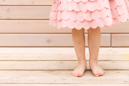 Beautifully beautiful legs of a child girl stand on a wooden terrace at home. Little girl in a pink fashionable dress barefoot on the street on a summer day. Outdoor partyの写真素材