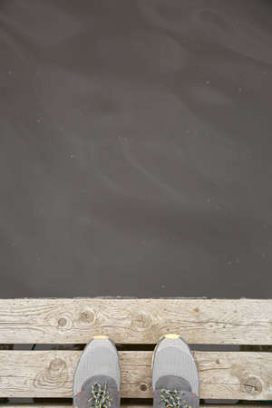 Top view on legs in sneakers standing on the edge of the pier. River, lake or just a pond in the...