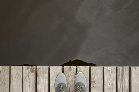 Top view on legs in sneakers standing on the edge of the pier. River, lake or just a pond in the...