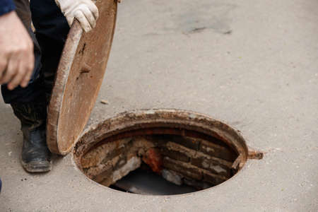 Dangerous open sewer manhole, a danger to people walking along the street within the city. Foreman closes the hatch cover. Repair of the pipeline under the ground.の写真素材