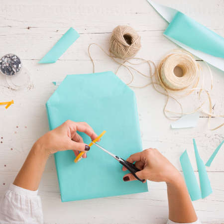 Close-up on a gift wrapped in paper of turquoise color. Hands of a young woman packing a gift on the background of a white wooden table. Preparations for the holiday. Accessories for packagingの写真素材