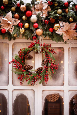 Festive Christmas wreath of evergreen spruce decorated with cones and red berries on a background of a garland with Christmas toys and a frozen window. Holiday concept.の写真素材