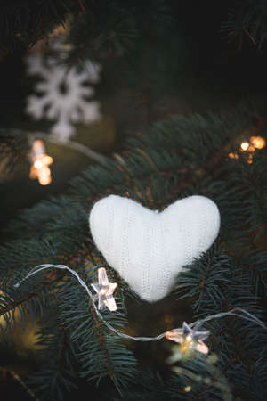 A soft knitted heart lies on the branches of a decorated Christmas tree on the background of Christmas decorations and a sparkling garland. Snowflake and star in the background. Holiday seasonの写真素材
