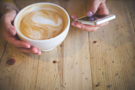 A young girl with a phone in her hands sits at a wooden table and types text using the touch screen of the phone. Top view of a girl with a phone and a cup of latte coffee. Communicationの写真素材