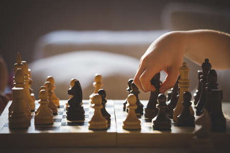 Cute boy sits at a table and plays chess. The kid picked up the chess piece of the elephant and makes his move. Early development, home educational games for children. Toning and artificial noiseの写真素材