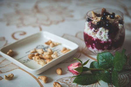 A bowl of organic cottage cheese yogurt with granola and black currant, a garden rose and a plate of cashew nuts on the table. Healthy eating concept. Toning and artificial noiseの写真素材