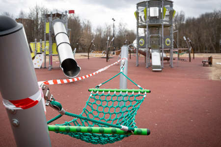 Empty playground with swings in a city park, fenced with warning tapes of red and white. Coronavirus pandemic covid-19 in the world. Stay at homeの写真素材