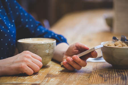A young girl with a phone in her hands sits at a wooden table and types text using the touch screen...