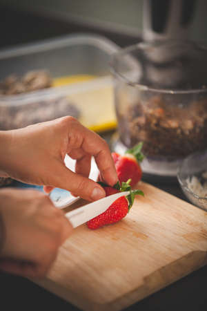 Girl prepares treats on the table by slicing strawberries. Healthy sweets. Organic raw treats from nuts, coconut, dates and cocoa and strawberries. Toning. Directly aboveの写真素材