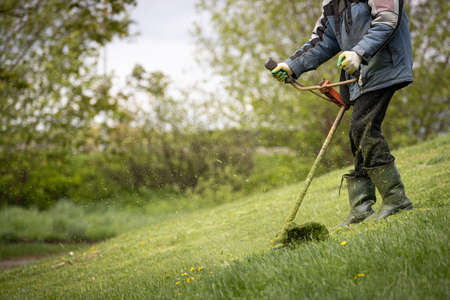 A man with a lawn mower mows grass and daisies on a slope near the house on a sunny spring day. A man in protective clothing, gloves and rubber boots mows the grass with a trimmer.の写真素材