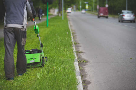 A worker in protective clothing and gloves with a gasoline mower on wheels walks along the lawn along the roadway. A man mows grass with dandelions next to passing cars in the city.の写真素材