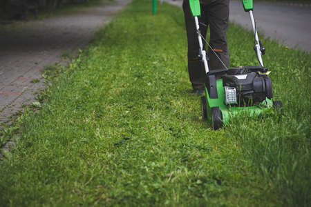 Front view of a worker in protective clothing with a gasoline lawn mower on wheels walking along the lawn along the roadway. A man mows grass with dandelions next to a road in the city.の写真素材