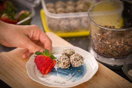 Top view on a plate with delicious homemade protein balls and ripe strawberries in the girl's hand. Healthy sweets. Organic raw treats from nuts, coconut, dates and cocoaの写真素材