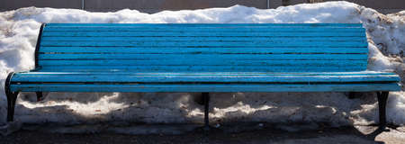 A bright blue, old wooden bench outside in the city on a warm spring day. Painted wood. The beginning of spring. Melting snow drifts. Some rubbish under the bench. Wide viewの写真素材