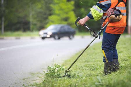 A worker in special reflective clothing with a gasoline mower in his hand. A man with a trimmer mows...