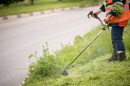 A worker in special reflective clothing with a gasoline mower in his hand. A man with a trimmer mows grass with dandelions on the side of the roadの写真素材