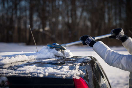 A girl with a brush in her hands cleans the roof of the car from snow. The car is covered with a...