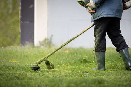 A man with a lawn mower on a grassy field near the house. A man in protective clothing, seals and rubber boots mows grass with a trimmer.の写真素材