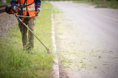 Worker in special protective reflective clothing with a lawn mower in his hands, mows grass with...