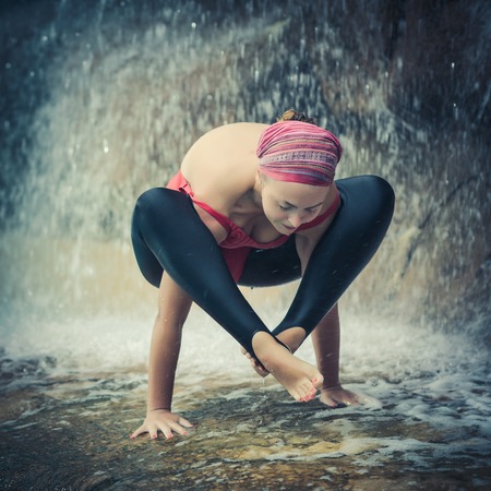 Woman practicing yoga near waterfall. Shoulder pressing posture. Bhujapidasanaの写真素材