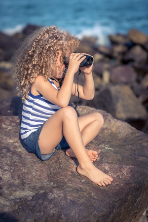 Little girl looking far away with binoculars, sitting on a rock near a seaの写真素材