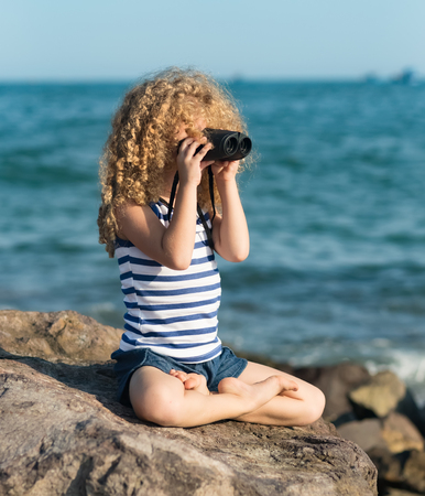 Little girl looking far away with binoculars, sitting on a rock near a seaの写真素材