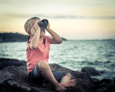 Little boy looking far away with binoculars, sitting on a rock near a seaの写真素材