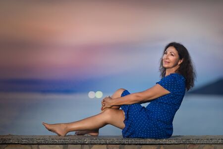 Outdoor portrait of a beautiful smiling senior woman sitting near the sea at sunsetの写真素材