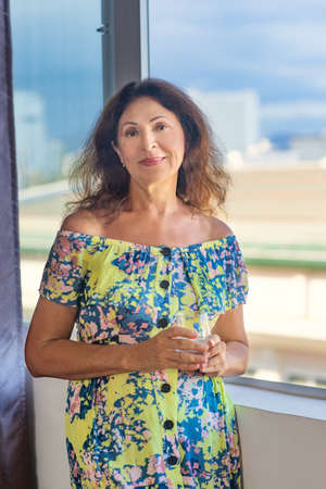 Indoor portrait of a beautiful smiling senior woman next to window holding glass of waterの写真素材