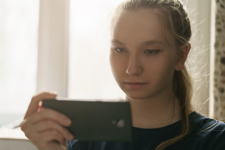 teen girl sitting near window with smartphone, toned photo with shallow focusの写真素材