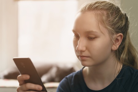 teen girl sitting near window with smartphone, toned photo with shallow focusの写真素材