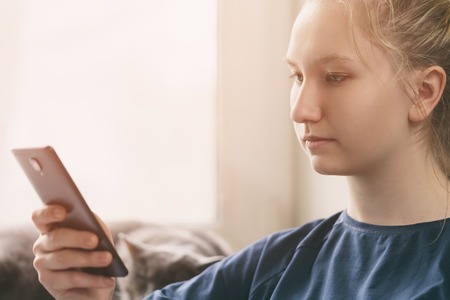 teen girl sitting near window with smartphone, toned photo with shallow focusの写真素材
