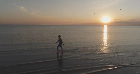 Aerial shot over teen girl barefoot walking in water on baltic sea beach in sunset time, wide photoの写真素材