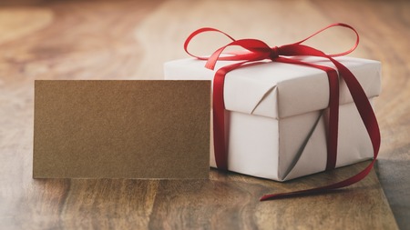 white gift box with thin red ribbon bow on old wood table with paper cardの写真素材