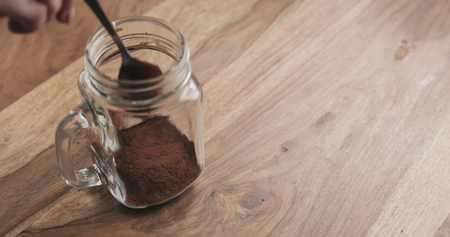 making cocoa drink in glass jar with handle on wood table, adding cacao powderの写真素材