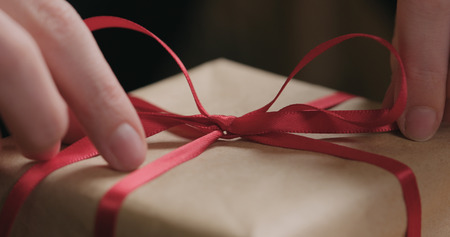 closeup shot of young female hands unties red ribbon bow on craft paper gift box, wide photoの写真素材